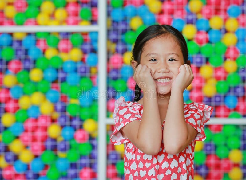 Den lyckliga lilla asiatiska barnflickan h?ller b?da h?nder p? kinder mot f?rgrik bolllekplats Charmigt och positivt se royaltyfria foton Den lyckliga lilla asiatiska barnflickan h?ller b?da h?nder p? kinder mot f?rgrik bolllekplats Charmigt och positivt se royaltyfria foton