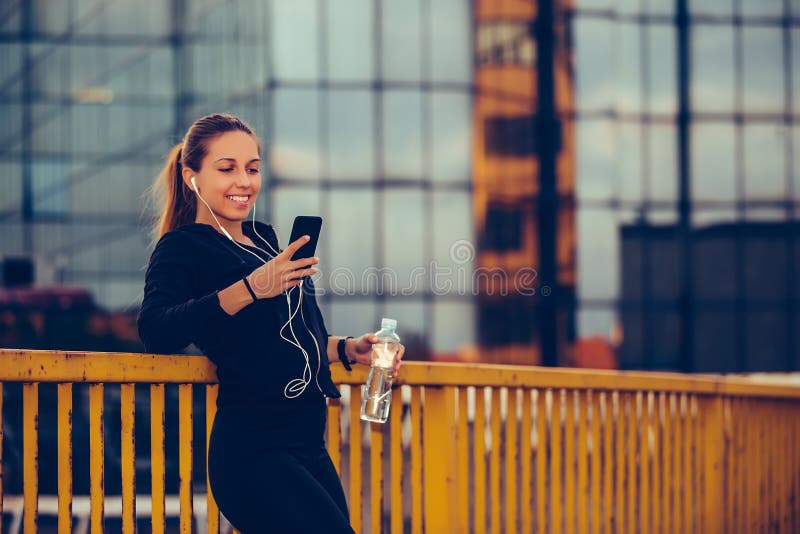 Den lyckliga sportiga flickan som tar ett avbrott efter, utarbetar och lyssnande musik, medan genom att använda den smarta telefo royaltyfri fotografi Den lyckliga sportiga flickan som tar ett avbrott efter, utarbetar och lyssnande musik, medan genom att använda den smarta telefo royaltyfri fotografi