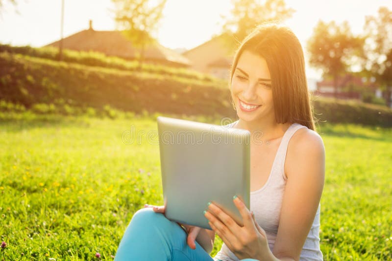 Happy beautiful young Caucasian brunette woman with tablet in park on sunny summer day. Back lit image. Modern lifestyle concept. Happy beautiful young Caucasian brunette woman with tablet in park on sunny summer day. Back lit image. Modern lifestyle concept.