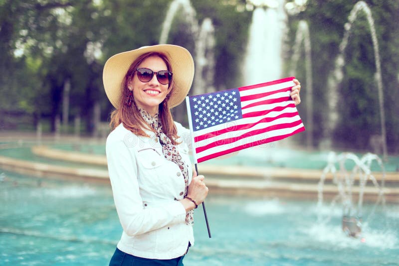 Den lyckliga unga patriotkvinnan i hatten som sträcker USA flaggan parkerar in royaltyfria foton Den lyckliga unga patriotkvinnan i hatten som sträcker USA flaggan parkerar in royaltyfria foton