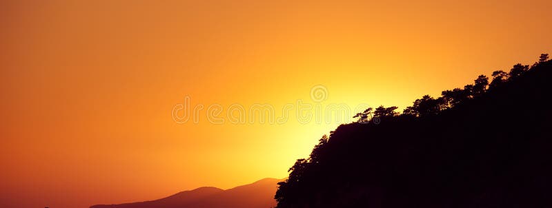 Den mörka konturn av ett berg som är bevuxet med träd mot den orange aftonhimlen Orange solnedg?ngbakgrund royaltyfri foto Den mörka konturn av ett berg som är bevuxet med träd mot den orange aftonhimlen Orange solnedg?ngbakgrund royaltyfri foto