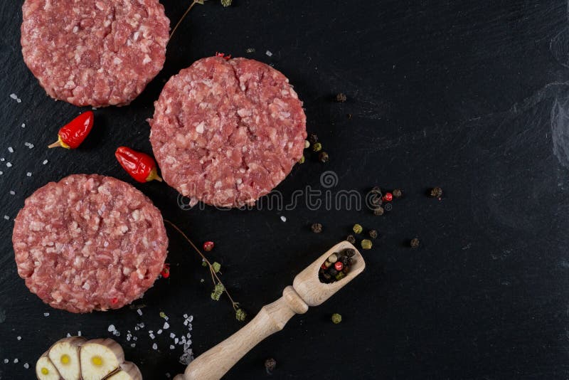 Den nya hamburgarekotletten för rått kött på svarten kritiserar brädet med örter och kryddor för bakgrund royaltyfri foto Den nya hamburgarekotletten för rått kött på svarten kritiserar brädet med örter och kryddor för bakgrund royaltyfri foto