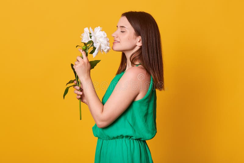Den nära övre profilståenden av damen som bär gröna sundress, håller härliga blommor i händer på gul studiobakgrund och att vara royaltyfria bilder Den nära övre profilståenden av damen som bär gröna sundress, håller härliga blommor i händer på gul studiobakgrund och att vara royaltyfria bilder