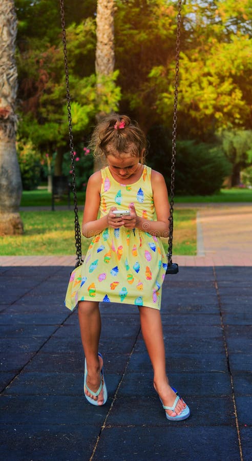 Den nätta unga flickan som spelar smartphonesammanträde på kedja, svänger på ungelekplatsen i parkera All hennes uppmärksamhet ti royaltyfri bild Den nätta unga flickan som spelar smartphonesammanträde på kedja, svänger på ungelekplatsen i parkera All hennes uppmärksamhet ti royaltyfri bild