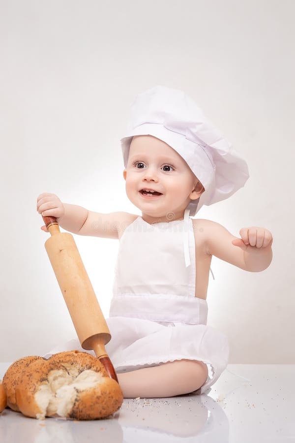 Den roliga kocken behandla som ett barn i kockdräkt- och kockhatten med degkavlen som sitter med bröd royaltyfri fotografi Den roliga kocken behandla som ett barn i kockdräkt- och kockhatten med degkavlen som sitter med bröd royaltyfri fotografi