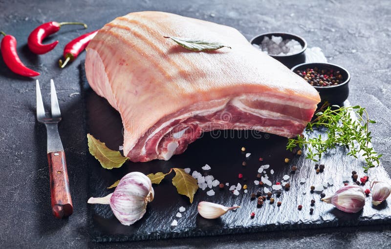 Raw pork belly with skin on a black slate tray with spices and herbs, horizontal view from above, close-up. Raw pork belly with skin on a black slate tray with spices and herbs, horizontal view from above, close-up
