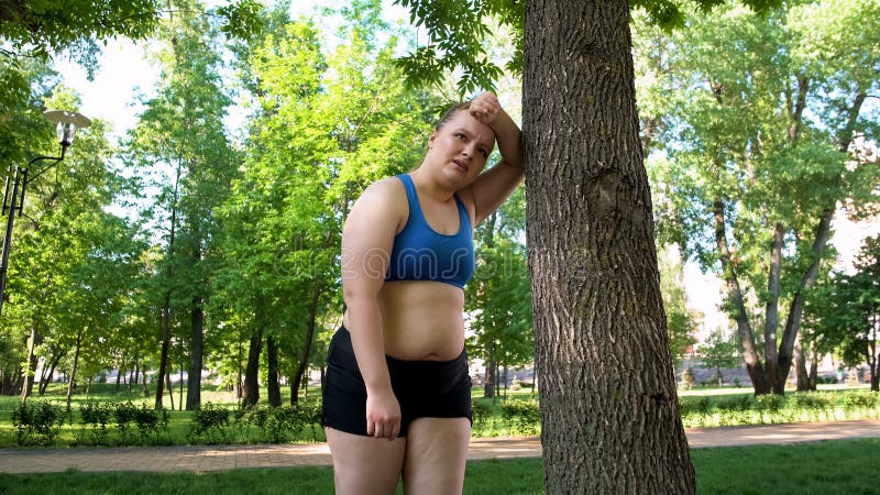 Den sjukligt feta kvinnan som ser trött efter genomkörare parkerar in och att luta på träd, motivationen royaltyfri foto Den sjukligt feta kvinnan som ser trött efter genomkörare parkerar in och att luta på träd, motivationen royaltyfri foto