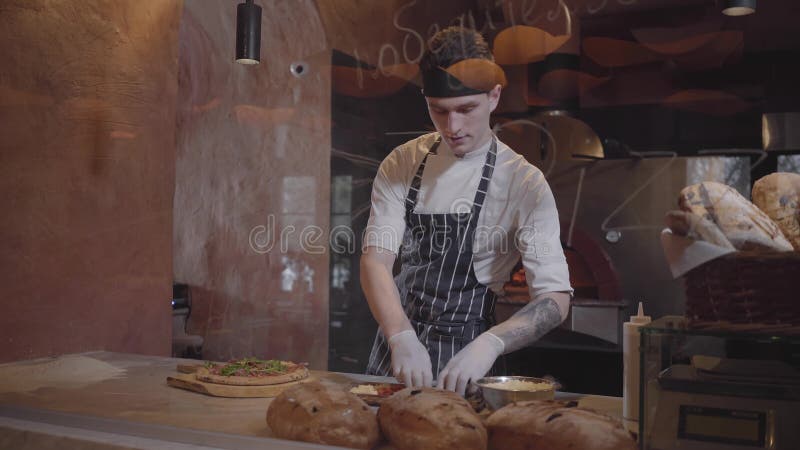 Den skickliga pizzaioloen lagar mat pizza på modernt restaurangkök bak exponeringsglas Ung grabb, i att sätta för kocklikformig