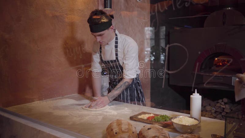 Den skickliga pizzatillverkaren lagar mat deg på modernt restaurangkök bak exponeringsglas Ung grabb i kocklikformign som rotera