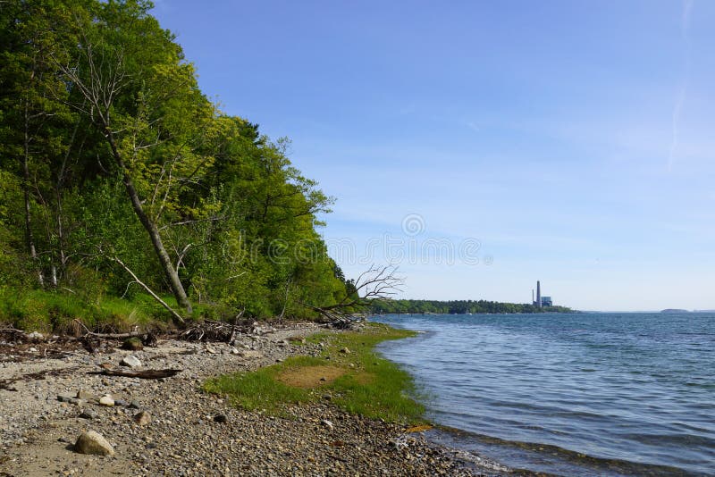 Rocky beach lined with trees on Cousins Island with Large Gas Power Plant in the distance in Yarmouth, Maine. Rocky beach lined with trees on Cousins Island with Large Gas Power Plant in the distance in Yarmouth, Maine.