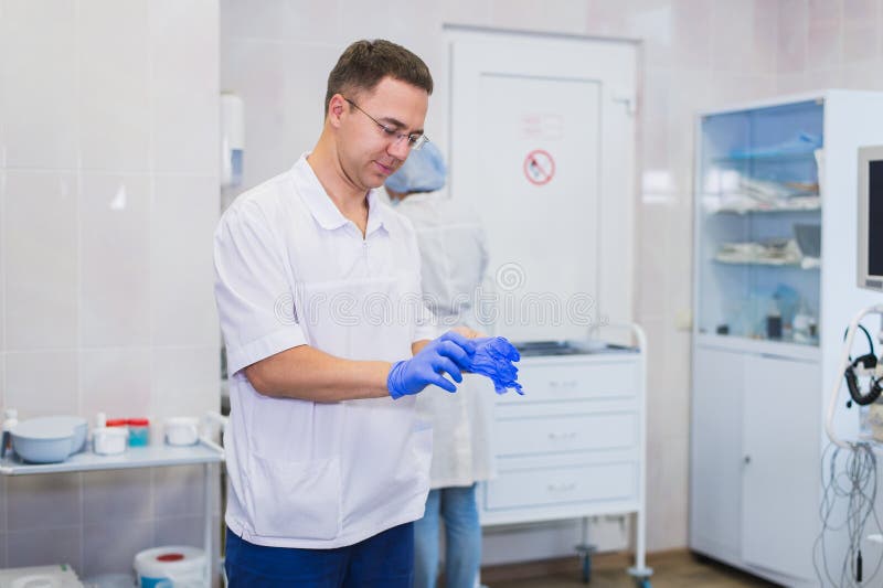 Den stiliga mogna kirurgen i blå medicinsk kläder och maskering sätter på medicinska handskar på fungeringsrum arkivbild Den stiliga mogna kirurgen i blå medicinsk kläder och maskering sätter på medicinska handskar på fungeringsrum arkivbild