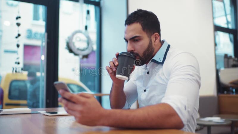 Den stiliga skäggiga mannen i en vit formell skjorta använder hans telefon för internet som surfar, medan dricka kaffe modernt lager videofilmer