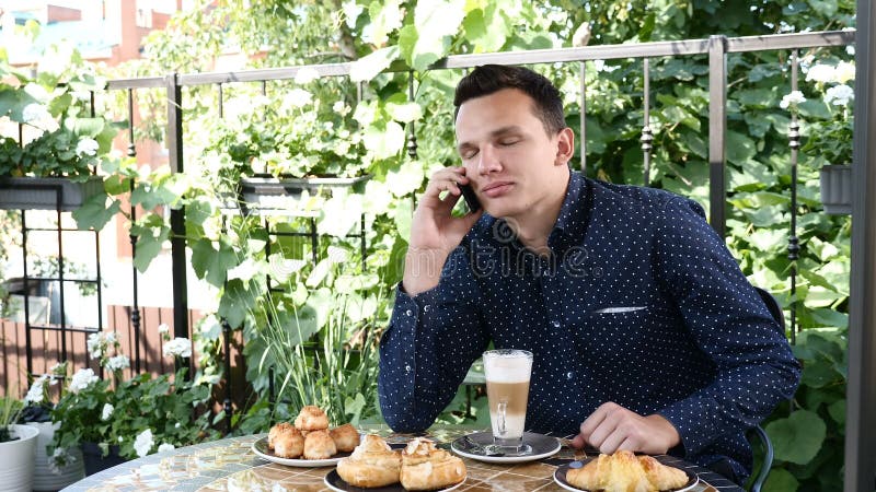 Den stiliga unga le mannen sitter i ett kafé som talar på telefonen som dricker cappuccino arkivfilmer