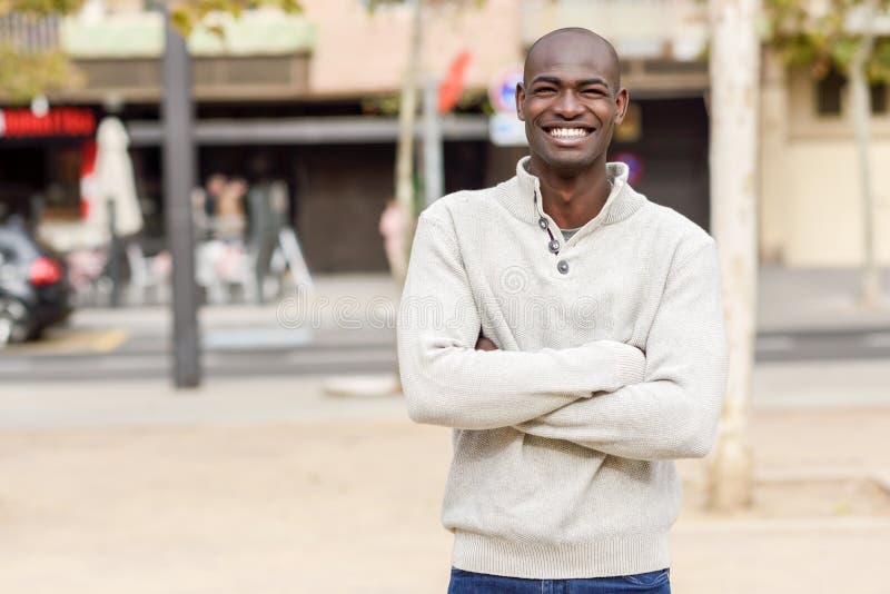 Black young man with arms crossed smiling in urban background. Young african guy with shaved head wearing casual clothes. Black young man with arms crossed smiling in urban background. Young african guy with shaved head wearing casual clothes.