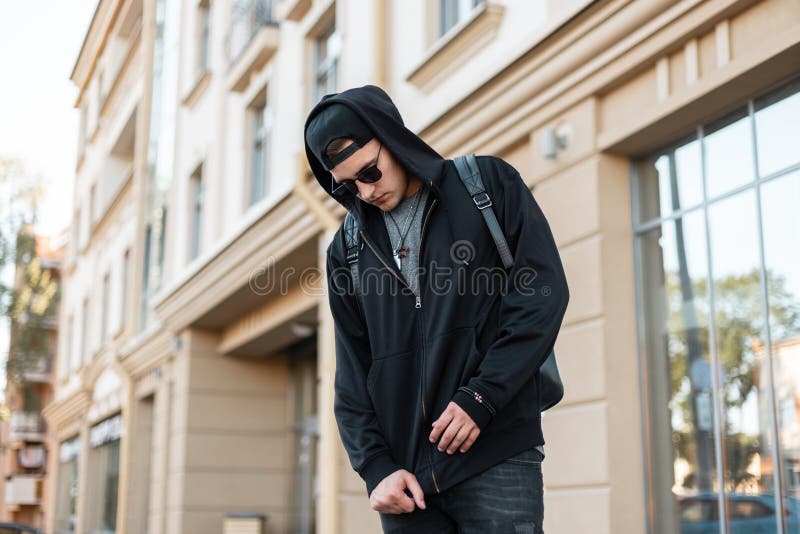 Den trevliga unga hipstermannen i en stilfull med huva tröja i moderiktig solglasögon i ett lock med en ryggsäck står i en stad royaltyfri bild Den trevliga unga hipstermannen i en stilfull med huva tröja i moderiktig solglasögon i ett lock med en ryggsäck står i en stad royaltyfri bild