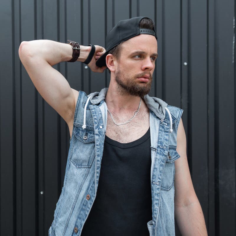 Den trevliga unga mannen som hipsteren med ett skägg i ett stilfullt svart lock i en trendig blå grov bomullstvill tilldelar en m royaltyfria bilder Den trevliga unga mannen som hipsteren med ett skägg i ett stilfullt svart lock i en trendig blå grov bomullstvill tilldelar en m royaltyfria bilder