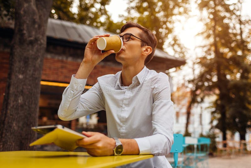 Den unga affärsmannen tycker om kaffe i utomhus- kafé genom att använda minnestavlan Stilig grabb som dricker te på stadsgatan arkivfoto Den unga affärsmannen tycker om kaffe i utomhus- kafé genom att använda minnestavlan Stilig grabb som dricker te på stadsgatan arkivfoto