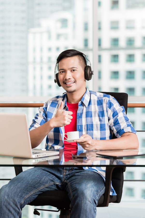 Den unga asiatiska mannen som har kaffe som ger tummar, up i regeringsställning tecknet royaltyfri bild Den unga asiatiska mannen som har kaffe som ger tummar, up i regeringsställning tecknet royaltyfri bild