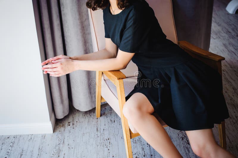 Den unga attraktiva kvinnan med långa ben i svart elegant klänning, sitter i stol nära fönster i inre av rum arkivfoton Den unga attraktiva kvinnan med långa ben i svart elegant klänning, sitter i stol nära fönster i inre av rum arkivfoton