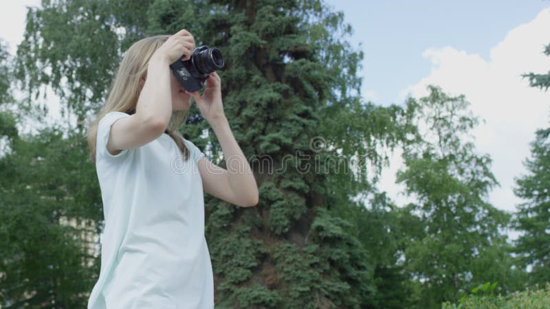 Den unga beskållade blonda kvinnan tar bilder i parken på sommardagen och lutar sig framåt och bakåt och letar efter de bästa