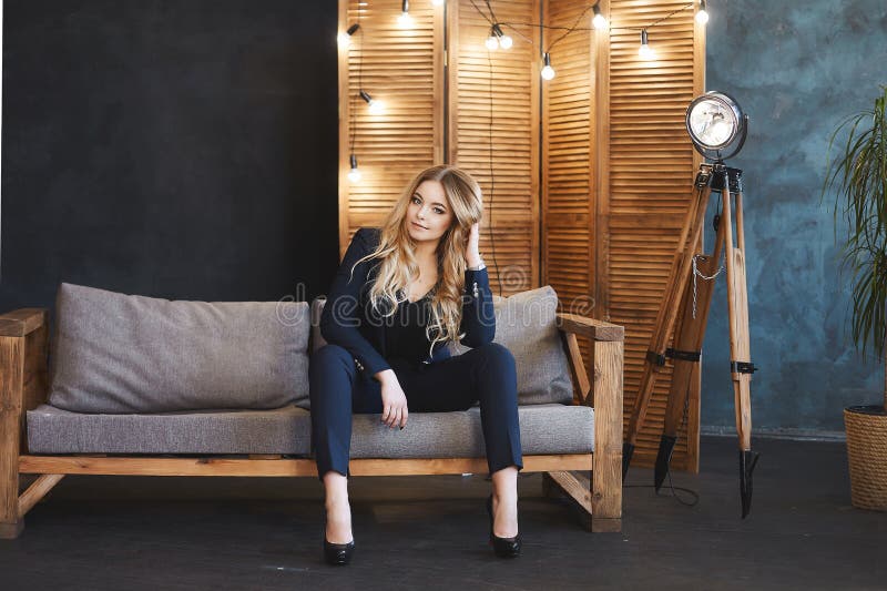 Den unga blonda affärsdamen i blåttdräkt sitter på träsoffan i modern inre royaltyfri fotografi Den unga blonda affärsdamen i blåttdräkt sitter på träsoffan i modern inre royaltyfri fotografi