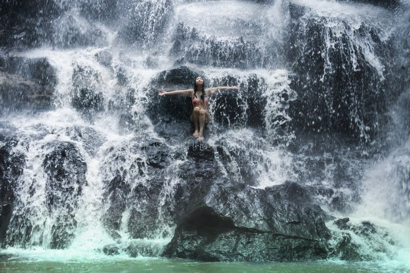 Den unga härliga och söta asiatiska kvinnan i bikinin som får kroppen den våta under-strömmen av naturligt fantastiskt vattenfall fotografering för bildbyråer