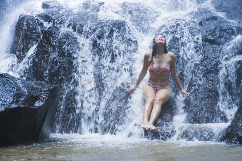 Den unga härliga och söta asiatiska kvinnan i bikinin som får kroppen den våta under-strömmen av naturligt fantastiskt vattenfall fotografering för bildbyråer