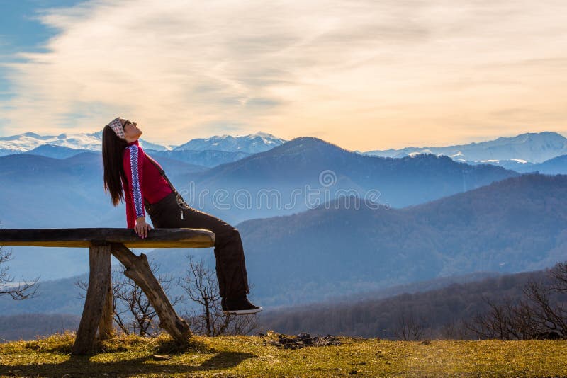 Den unga kvinnan sitter på bänk utanför och blick på det bildmässiga landskapet med berg royaltyfria foton Den unga kvinnan sitter på bänk utanför och blick på det bildmässiga landskapet med berg royaltyfria foton