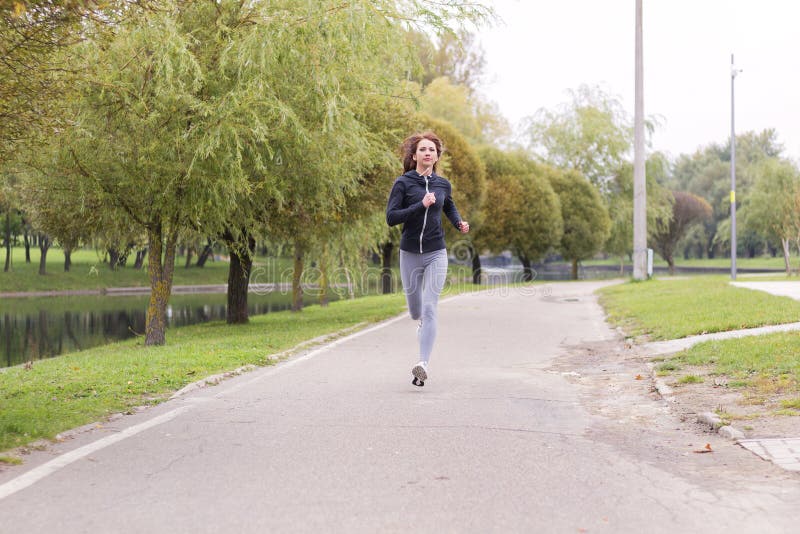 Den unga lyckliga kvinnan som joggar i höst, parkerar Rinnande konditionflicka royaltyfri fotografi Den unga lyckliga kvinnan som joggar i höst, parkerar Rinnande konditionflicka royaltyfri fotografi