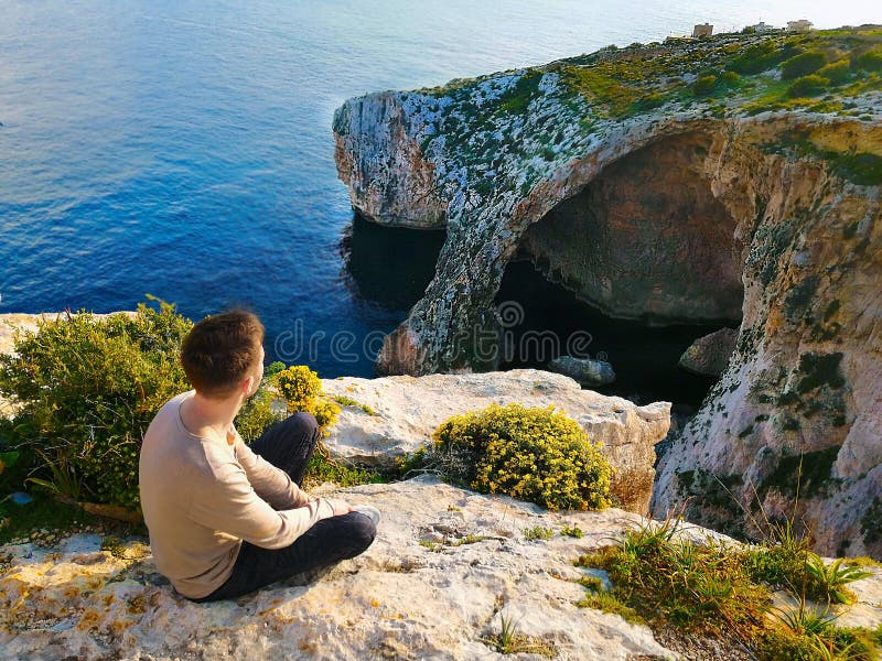 Den unga mannen sitter på kanten av en klippa och beundrar havet och stenarna royaltyfria foton Den unga mannen sitter på kanten av en klippa och beundrar havet och stenarna royaltyfria foton