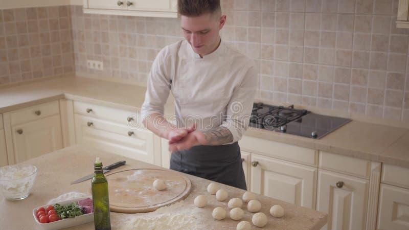 Den unga pizzatillverkaren i kocklikformig häller olivolja förestående som rullar degbollar för pizza i köket kvinna för vektor f