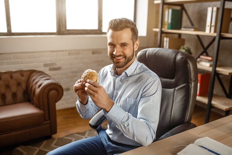 Den unga stiliga affärsmannen sitter på stol och att ha lunchtid i hans eget kontor Han rymmer hamburgaren och leende till kamera royaltyfri bild Den unga stiliga affärsmannen sitter på stol och att ha lunchtid i hans eget kontor Han rymmer hamburgaren och leende till kamera royaltyfri bild