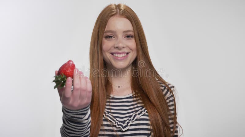 Den unga söta ljust rödbrun flickan visar jordgubben och att le, vit bakgrund