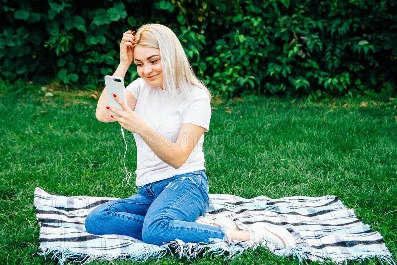 Den vackra blonda kvinnan i hörlurarna och med en smartphone i händerna, titta på apparaten som sitter på gräs utomhus i parken arkivbilder