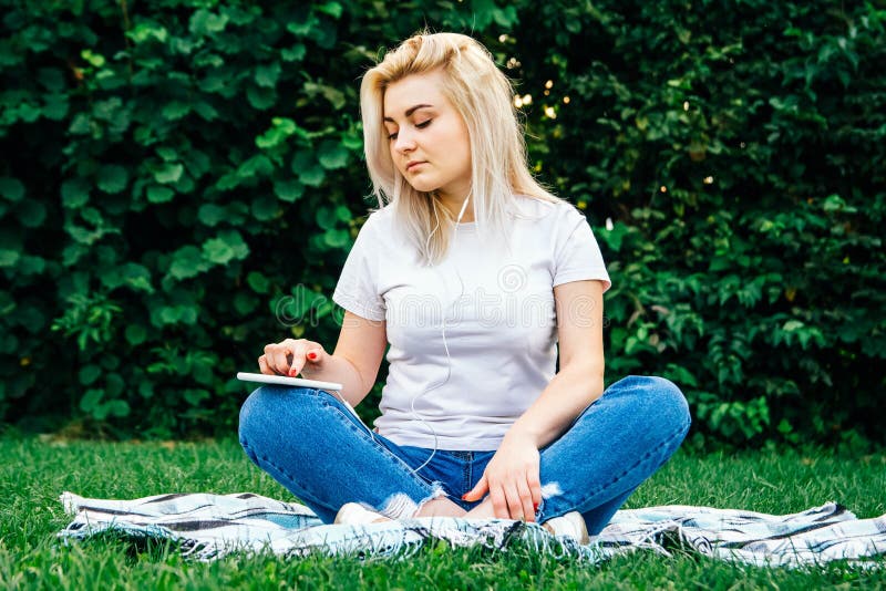 Den vackra blonda kvinnan i hörlurarna och med en smartphone i händerna, titta på apparaten som sitter på gräs utomhus i parken royaltyfria foton