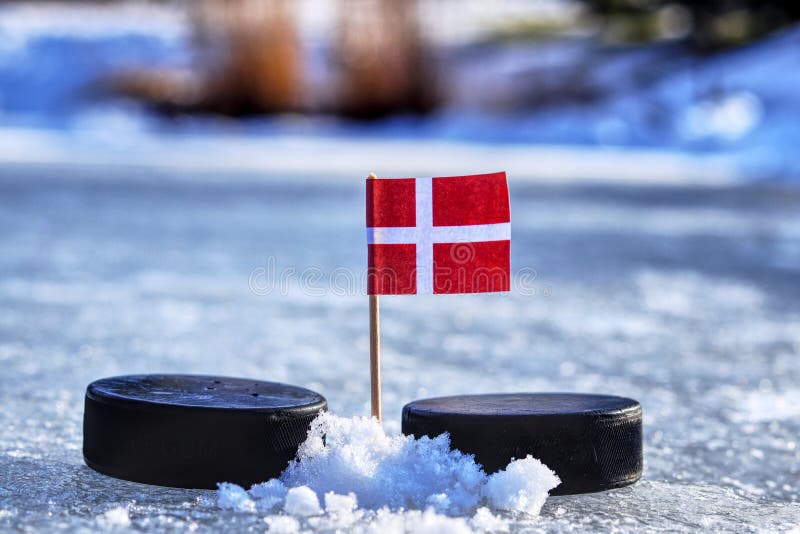 Dánská vlajka na párátko mezi dvěma hokejovými puky. Dánsko bude hrát na mistrovství světa ve skupině A. 2019 IIHF World Championship