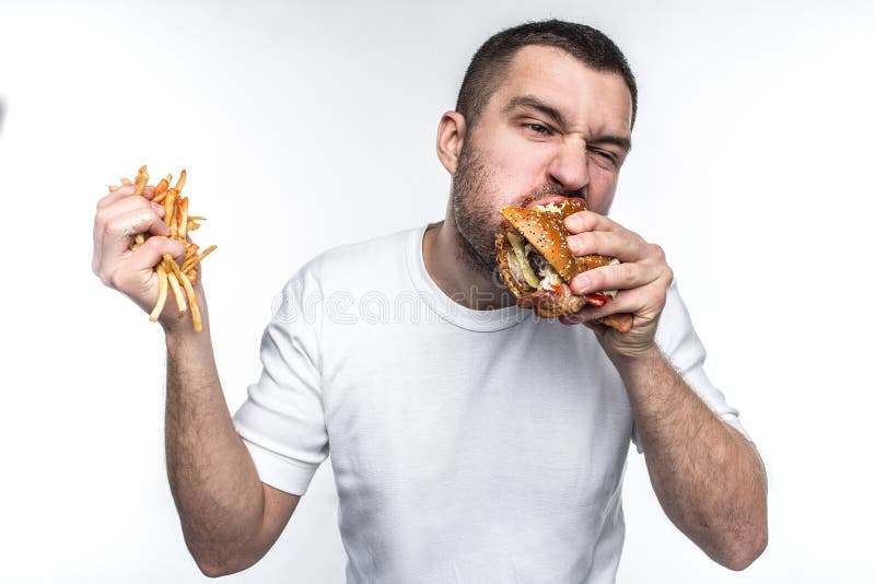 This guy is very delight of junk food. He is biting a big piece of burger and holding a full hand of french fries covered with ketchup. Young amn likes to eat oily meal. Isolated on white background. This guy is very delight of junk food. He is biting a big piece of burger and holding a full hand of french fries covered with ketchup. Young amn likes to eat oily meal. Isolated on white background