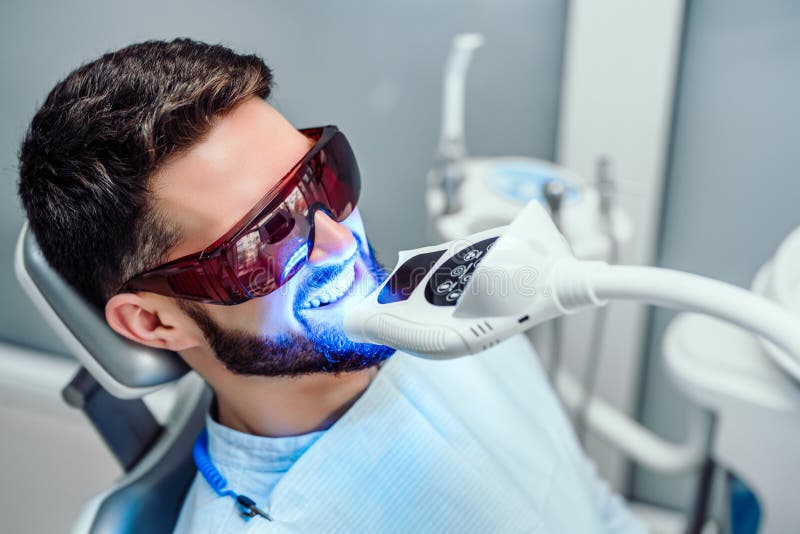 Dentista que enciende los dientes el blanquear de procedimiento con el hombre joven
