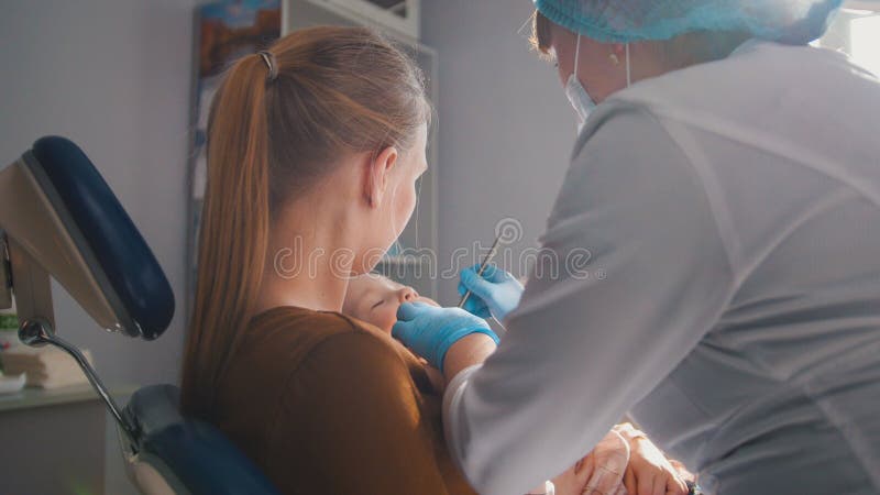 Dentista que examina e que consulta a filha que senta-se em seus joelhos da mãe imagem de stock royalty free Dentista que examina e que consulta a filha que senta-se em seus joelhos da mãe imagem de stock royalty free