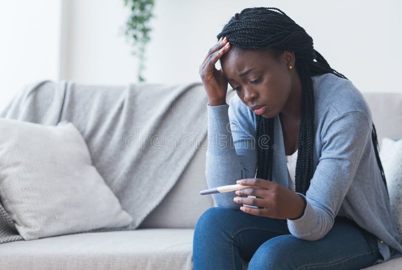 Unintended pregnancy. Depressed african american woman upset with positive test results, sitting on couch at home with sad face expression. Unintended pregnancy. Depressed african american woman upset with positive test results, sitting on couch at home with sad face expression