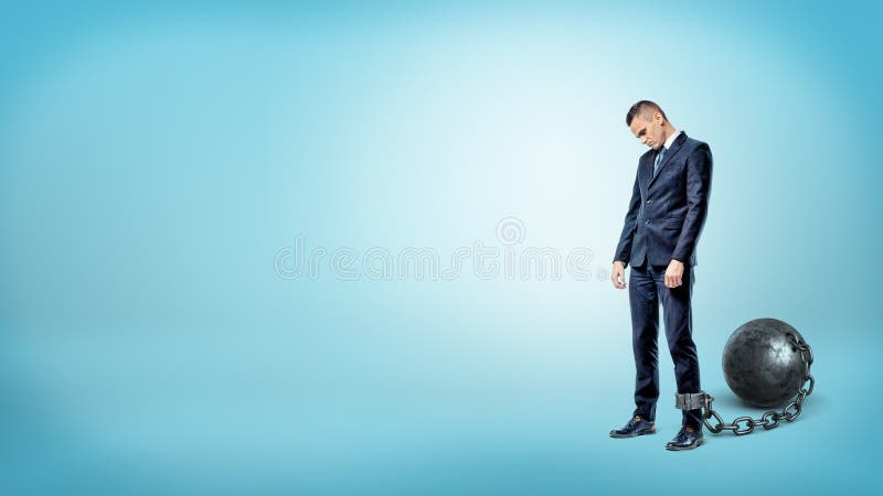 A depressed businessman on blue background stands with a lowered head while chained to an iron ball. stock photo A depressed businessman on blue background stands with a lowered head while chained to an iron ball. stock photo
