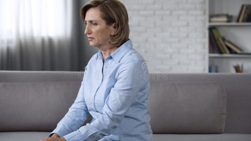 Depressed senior lady sitting on couch, feeling anxious, psychological problems, stock photo