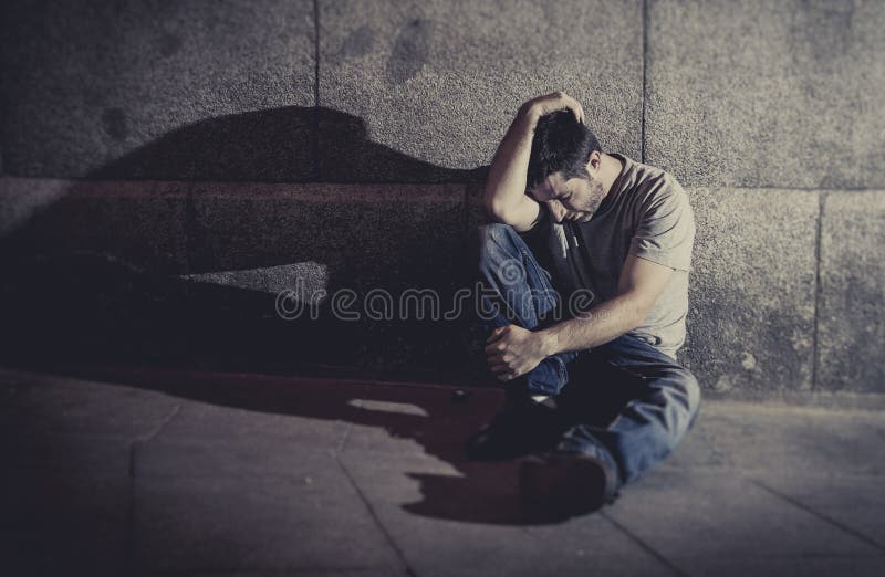 Depressed young man sitting on street ground with shadow on concrete wall
