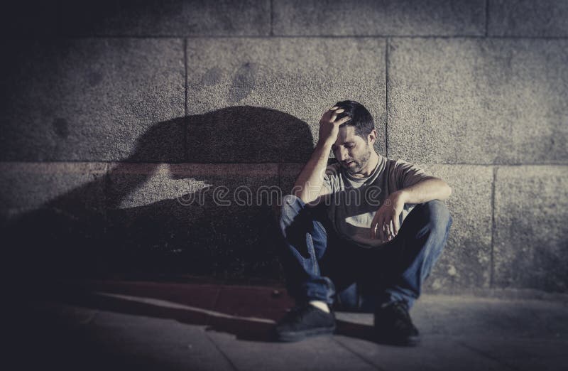 Depressed young man sitting on street ground with shadow on concrete wall