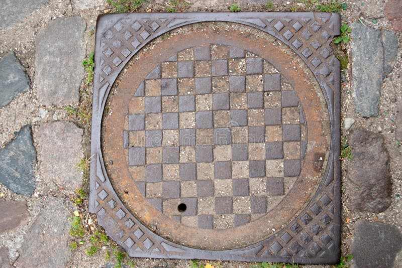 Der Brunnen der städtischen Abwasserkanalisation mit einem aus Metall gewellten, braunen Deckel mit Schachbrettmuster, unter den lizenzfreies stockfoto