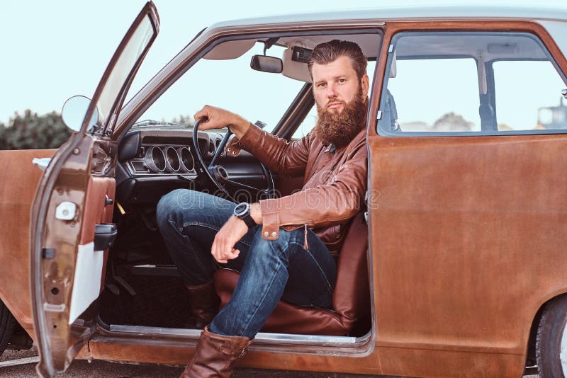 Stylish bearded male dressed in brown leather jacket sits behind the wheel of a tuned retro car with open door. Stylish bearded male dressed in brown leather jacket sits behind the wheel of a tuned retro car with open door.