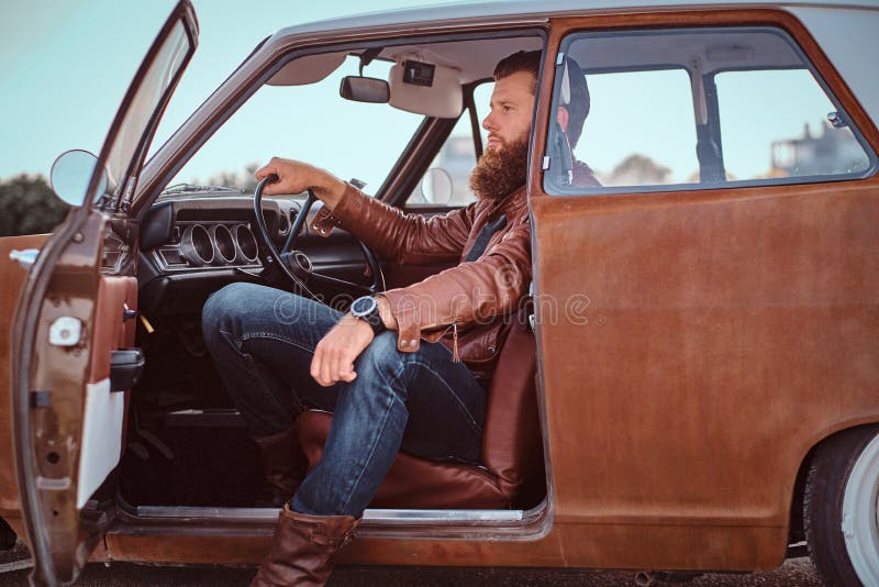 Stylish bearded male dressed in brown leather jacket sits behind the wheel of a tuned retro car with open door. Stylish bearded male dressed in brown leather jacket sits behind the wheel of a tuned retro car with open door.