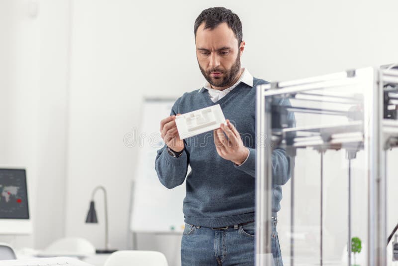 Careful inspection. Pleasant young man standing in the office and looking at the model printed on a 3D printer, carefully examining its quality. Careful inspection. Pleasant young man standing in the office and looking at the model printed on a 3D printer, carefully examining its quality