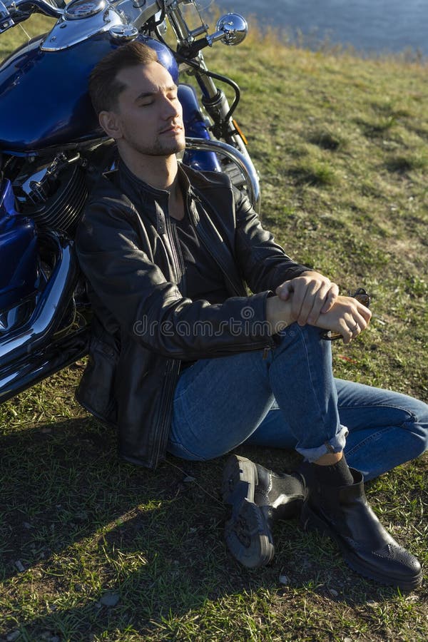 Der junge Mann, der eine schwarze Lederjacke und Jeans trägt, sitzt im Freien lizenzfreie stockbilder Der junge Mann, der eine schwarze Lederjacke und Jeans trägt, sitzt im Freien lizenzfreie stockbilder
