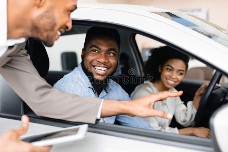 Happy Black Couple Buying New Car Testing And Talking With Salesman Sitting In Auto Dealership Showroom. Family Choosing Automobile. Happy Black Couple Buying New Car Testing And Talking With Salesman Sitting In Auto Dealership Showroom. Family Choosing Automobile.